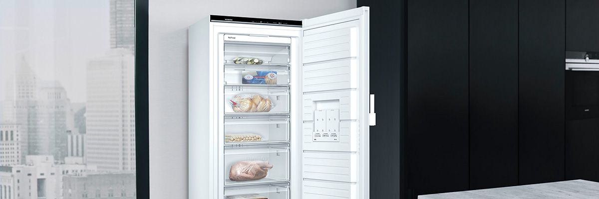 Weißer Kühlschrank in einem modernen Raum, offen mit Lebensmitteln gefüllt, vor schwarzen Wänden und Fenster mit Stadtblick.